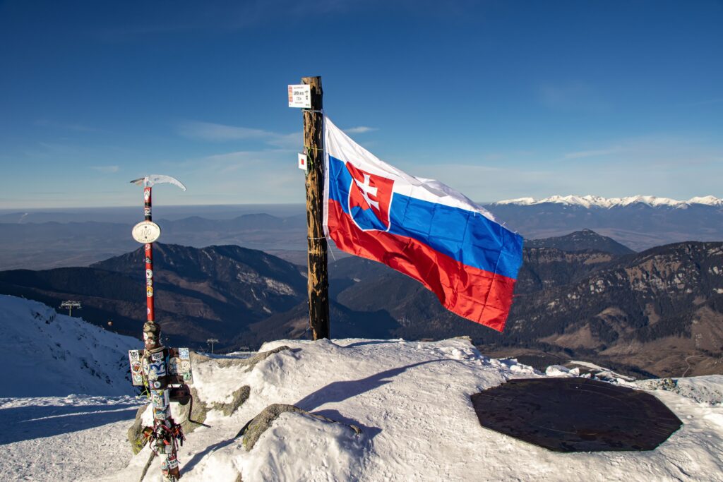 Slovak flag atop a snowy mountain with a panoramic view of distant ranges.